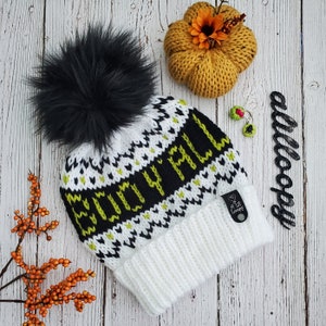 May include: White knit beanie with a black and green "Cozy Fall" design and a black faux fur pom pom. The beanie has a white knit cuff and a small black label with the text "A Lil Loopy" on it.