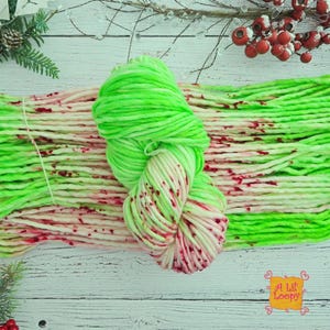 May include: A skein of thick, neon green yarn featuring a variegated pattern with splatters of bright red and white. The yarn is arranged in a horizontal bundle, showcasing a soft, chunky texture with a consistent color distribution throughout.