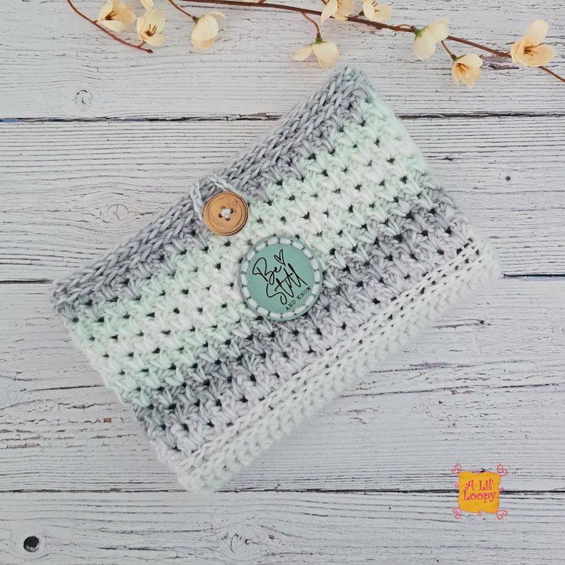 Pode incluir: Um aquecedor de pesco&ccedil;o de croch&ecirc; em tons de cinza, branco e verde menta, com fecho de bot&atilde;o de madeira e um patch circular com as palavras "Be Still And Know". O cachecol feito &agrave; m&atilde;o &eacute; um acess&oacute;rio aconchegante.