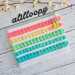 May include: A rainbow crochet pouch with a white background and a fluffy texture. The pouch has a small silver key chain with two yellow tassels attached.
