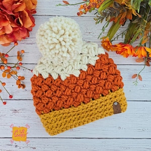 May include: A handmade crochet beanie in the colors of candy corn: cream, orange, and yellow. The hat features a large cream-colored pom-pom on top and a small leather tag. The text "A Lil' Loopy" is visible.