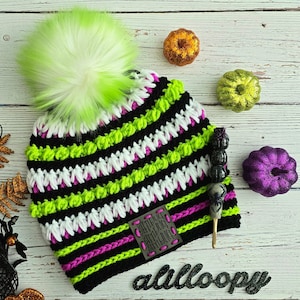 May include: A black, white, green, and pink crocheted hat with a fluffy white pom pom. The hat has a striped pattern and a black patch with the text "Brittney Jack Betelgeuse Betelgeuse Betelgeuse" on it. The hat is sitting on a white wooden surface with a crochet hook and some decorative pumpkins.