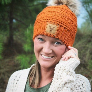 May include: A bright orange crocheted beanie with a faux fur pom pom and a brown leather patch featuring a deer silhouette. The beanie is being worn by a person with long brown hair and a white sweater.
