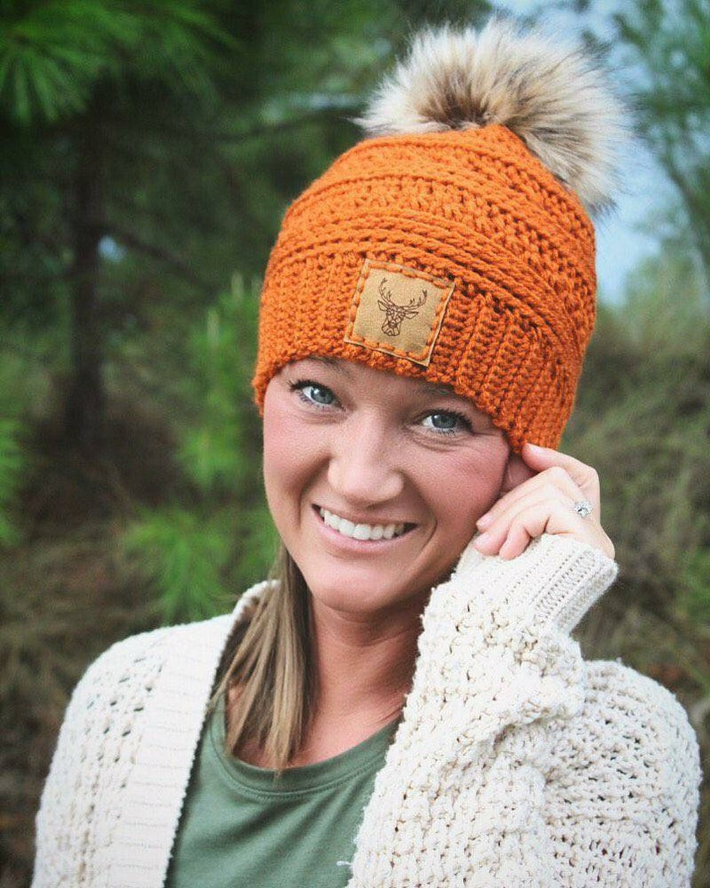May include: A bright orange crocheted beanie with a faux fur pom pom and a brown leather patch featuring a deer silhouette. The beanie is being worn by a person with long brown hair and a white sweater.