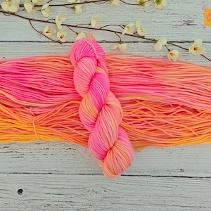 May include: A skein of hand-dyed yarn in vibrant shades of pink and orange, with a gradient effect. The yarn is arranged on a white wooden surface, with a sprig of white flowers in the background. The yarn is perfect for knitting or crocheting projects.