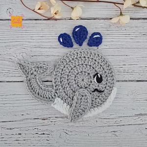 May include: A gray and white crocheted whale applique featuring a rounded body, a tail, and a side fin. Above the whale are three small, dark blue crocheted water droplets. The whale has a black embroidered eye and a small curved mouth.