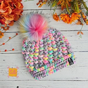 Peut inclure: Un bonnet crocheté à la main, gris et pastel, orné d'un gros pompon multicolore et duveteux. Le bonnet présente un motif de point texturé et une petite étiquette indiquant "A Lil' Loopy".