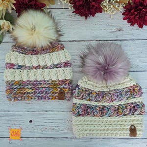 May include: Two crochet beanies featuring alternating bands of cream-coloured yarn and variegated pastel yarn in shades of pink, blue, and yellow. Each hat is topped with a large faux fur pom-pom and includes a small brown leather tag labelled A Lil Loopy.