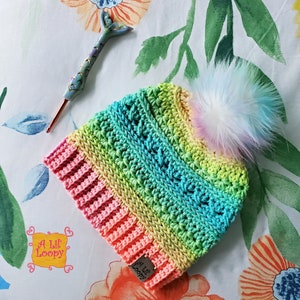May include: A rainbow crochet beanie with a fluffy white pom pom. The beanie has a pink, yellow, green, and blue colour gradient. The beanie has a tag that says "A Lil' Loopy".