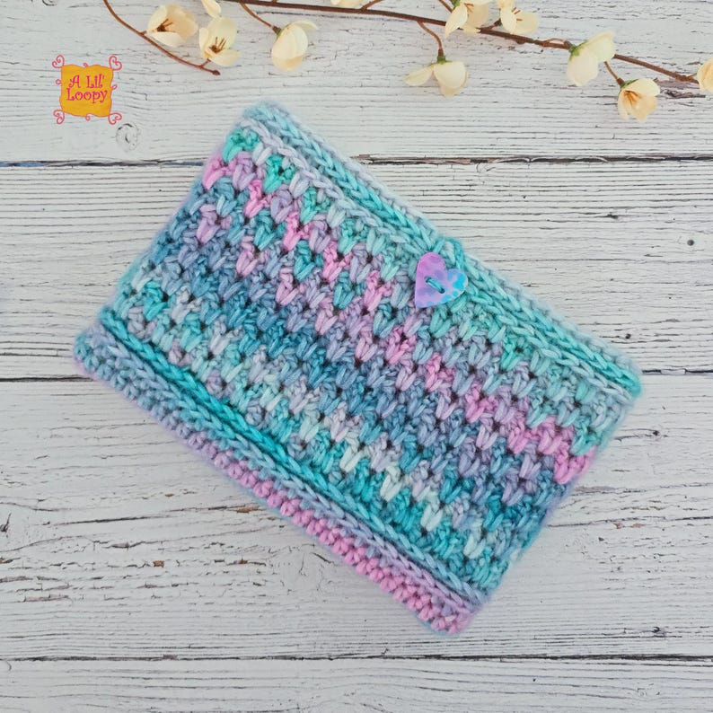 Pode incluir: Uma capa de tablet de croch&ecirc;, feita &agrave; m&atilde;o, em tons de azul, turquesa e rosa. Apresenta um ponto texturizado e um fecho de bot&atilde;o em forma de cora&ccedil;&atilde;o. A capa foi projetada para proteger um tablet. O texto "A Lil Loopy" &eacute; vis&iacute;vel.