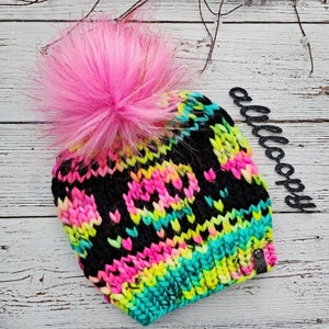 May include: A black, pink, yellow, and blue knit beanie with a fluffy pink pom pom on top. The beanie has a colorful, abstract pattern.