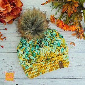 Puede incluir: Un gorro tejido a mano en tonos amarillos, turquesa y crema, rematado con un gran pompón de piel sintética. El gorro presenta un diseño de punto grueso y una pequeña etiqueta de cuero. El texto "A Lil' Loopy" es visible en la parte inferior izquierda.