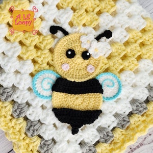 May include: A crocheted baby blanket with a yellow, white, and gray color scheme. The blanket features a crocheted bee applique with black and yellow stripes, blue wings, and a white flower on its head.