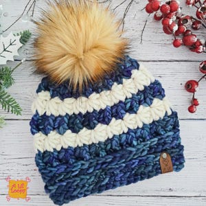Può includere: Un cappello invernale lavorato a mano con un grande pompon in finta pelliccia. Il cappello presenta strisce alternate di filato bianco e blu. Una piccola etichetta in pelle con la scritta "A Lil' Loopy" è attaccata.