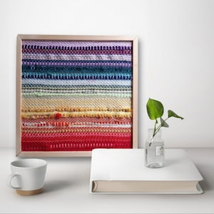May include: A framed wall hanging with a woven tapestry featuring a rainbow of colors in horizontal stripes. The tapestry is made of different colored yarns and is framed in a light wood frame.