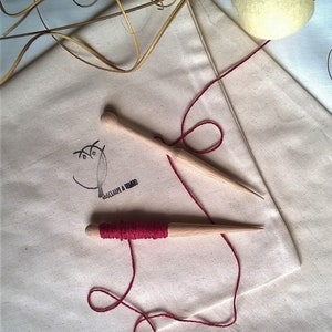 May include: Two wooden knitting needles with red yarn wrapped around one of them. The needles are on a white fabric surface with a brown owl logo.