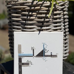 Op de afbeelding: Vierkante canvas kunst met een abstract ontwerp in tinten blauw, grijs en bruin. Het kunstwerk toont geometrische vormen en getextureerde lijnen. Een gevlochten mand en groen zijn op de achtergrond.