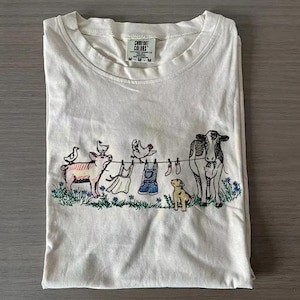 May include: Cream-coloured t-shirt featuring a farmyard scene. The design includes a pig, cow, dog, and chickens, with clothes hanging on a washing line. The shirt is folded and has a Comfort Colors label.