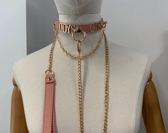 May include: Pink leather choker with gold lettering that reads "DADDYS GIRL". The choker has a gold ring and chain detail, with a matching leash. The leash and choker are displayed on a mannequin.