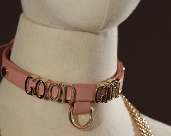May include: A pink collar with the words "GOOD GIRL" in gold lettering. The collar has a gold-colored D-ring and a gold chain attached. The collar is displayed on a mannequin.