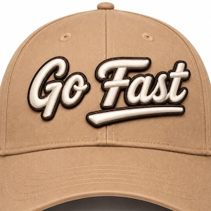 May include: A tan baseball cap featuring the text Go Fast in a raised, white, cursive font with a dark brown outline. The cap has a structured crown, a curved brim, and stitched eyelets on the top panels, presenting a clean, solid-colored design.