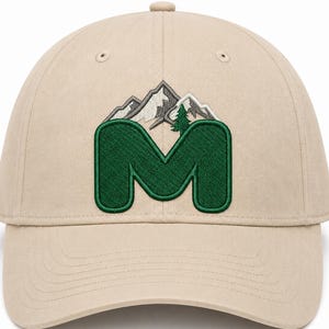 May include: A beige baseball cap featuring an embroidered design on the front. The design consists of a large, dark green letter M with a mountain range in shades of grey and white positioned behind it, along with a small green pine tree.