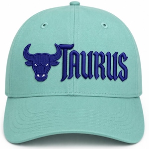 May include: A light mint green baseball cap featuring an embroidered dark blue bull head icon and the word TAURUS in a bold, stylized font. The cap has a structured crown with ventilation eyelets and a curved brim, showcasing detailed stitching.