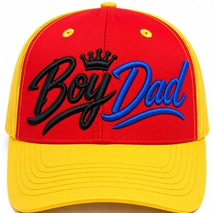 May include: A red and yellow baseball cap featuring the embroidered text "Boy Dad" in black and blue script, topped with a small black crown icon. The cap has a structured red front panel, a yellow brim with stitching, and yellow mesh side panels.