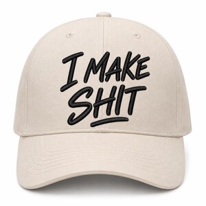 May include: A beige baseball cap featuring the black embroidered text I MAKE SHIT centered on the front panel. The cap has a structured crown, a curved brim, and a button at the top, constructed from a solid fabric with visible stitching.