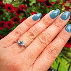 May include: A silver ring featuring a light blue gemstone set in a delicate, branch-like design. The ring is worn on the ring finger of a hand with blue nail polish.