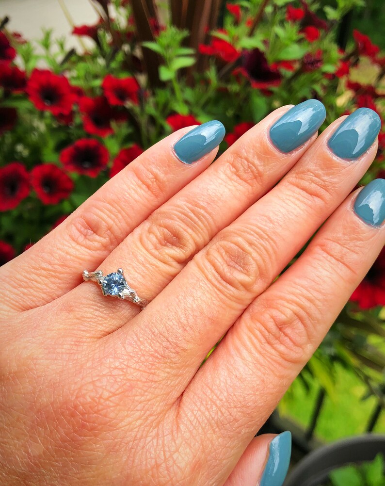 May include: A silver ring featuring a light blue gemstone set in a delicate, branch-like design. The ring is worn on the ring finger of a hand with blue nail polish.