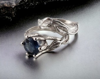 Puede incluir: Un anillo de plata con una piedra preciosa de zafiro azul. El anillo presenta un diseño delicado y frondoso con detalles intrincados. La banda está hecha de plata pulida y tiene un acabado liso.