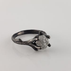 Puede incluir: Un anillo plateado con una gema en bruto de color gris claro. La banda y el engaste del anillo est&aacute;n dise&ntilde;ados para parecerse a una ramita, con delicadas ramas que se extienden para sostener la piedra. El anillo est&aacute; sobre un fondo blanco.