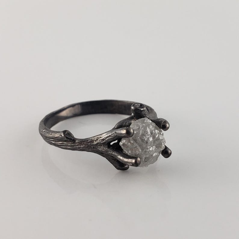 Puede incluir: Un anillo plateado con una gema en bruto de color gris claro. La banda y el engaste del anillo est&aacute;n dise&ntilde;ados para parecerse a una ramita, con delicadas ramas que se extienden para sostener la piedra. El anillo est&aacute; sobre un fondo blanco.