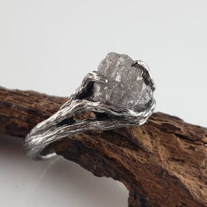 Op de afbeelding: Een zilveren ring met een ruwe, grijze edelsteen. De band en zetting van de ring hebben een textuur, organisch ontwerp, dat lijkt op verweven takken. De edelsteen staat centraal, wat een unieke en natuurlijke uitstraling cre&euml;ert.