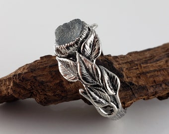 Puede incluir: Un anillo de plata con una piedra preciosa gris de corte rugoso engastada en un diseño detallado de hojas. La banda y el engaste del anillo están diseñados para parecerse a hojas delicadas, creando una estética inspirada en la naturaleza. El anillo se muestra sobre un trozo de madera.