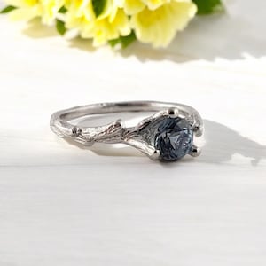May include: A silver ring featuring a branch design and a dark blue gemstone. The ring is displayed on a white wooden surface.