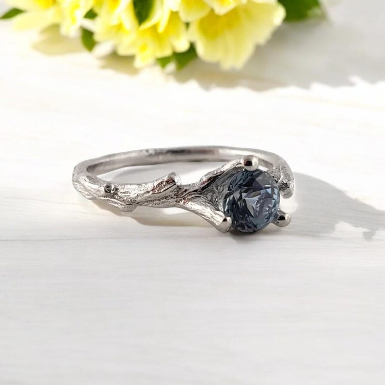 May include: A silver ring featuring a branch design and a dark blue gemstone. The ring is displayed on a white wooden surface.