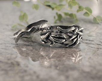 Puede incluir: Dos anillos de plata con un diseño de hojas. Los anillos están apilados uno encima del otro y están sobre una superficie gris.