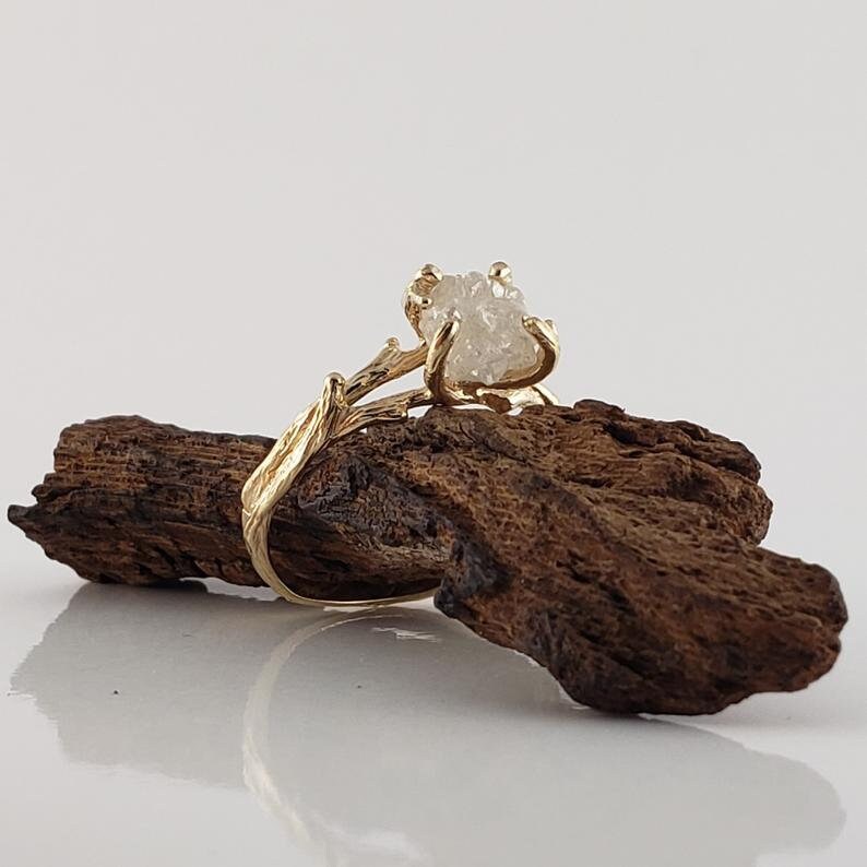 May include: A gold ring with a rough, white diamond set in a branch-like design. The ring is resting on a piece of dark brown driftwood.