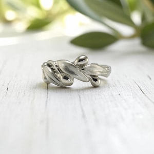 May include: A silver-colored ring with a floral design. The ring features a cluster of petal-like shapes and small, round accents. The ring is set against a blurred background of green foliage and a white wooden surface.