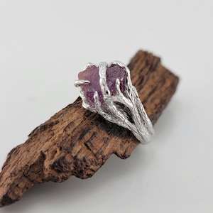 May include: A silver ring featuring a pink gemstone set within a tree branch design.