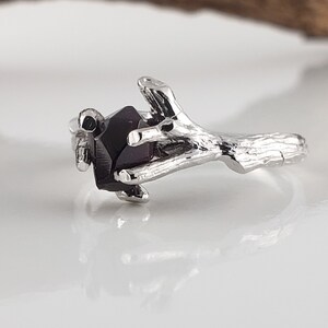 May include: A silver ring with a branch design holding a dark purple gemstone. The ring is made of sterling silver and features a unique and intricate design.