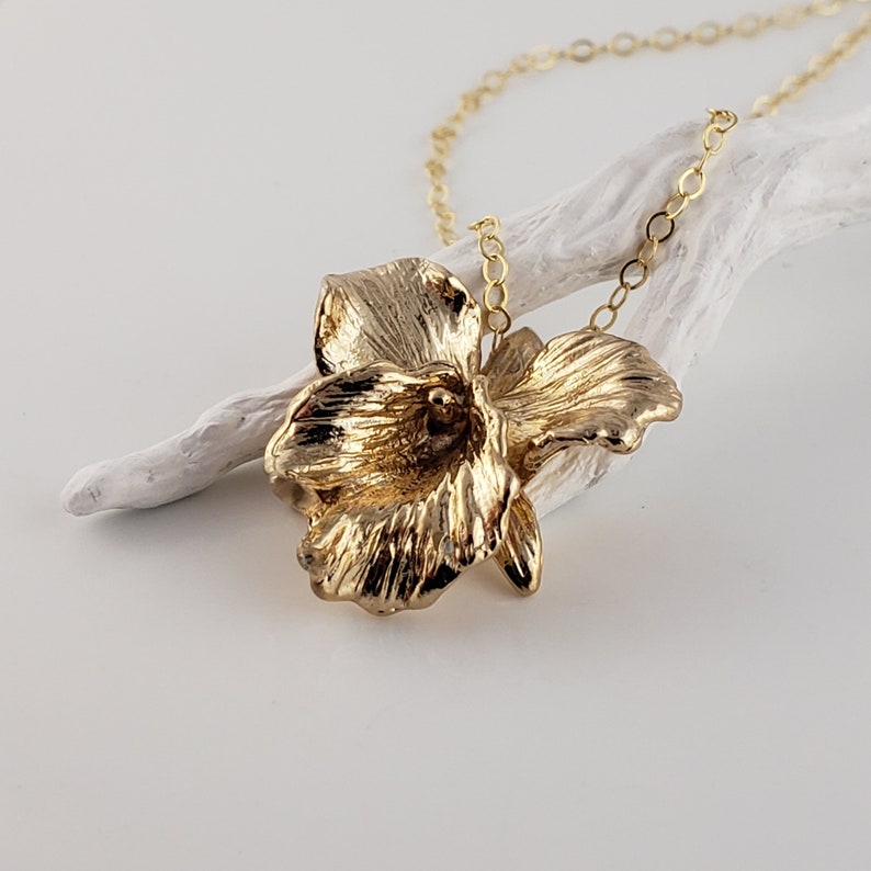 May include: A gold orchid flower pendant necklace. The pendant is detailed and textured, with a realistic orchid flower design. The necklace is made of a delicate gold chain.