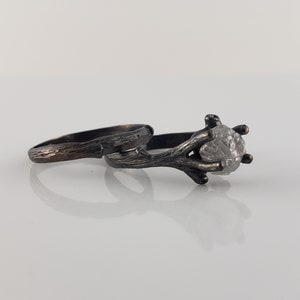 Puede incluir: Dos anillos de plata. Uno presenta un diamante en bruto de color gris claro sujeto por un engaste en forma de ramita. El otro anillo tiene una banda texturizada. Ambos anillos est&aacute;n sobre un fondo blanco.