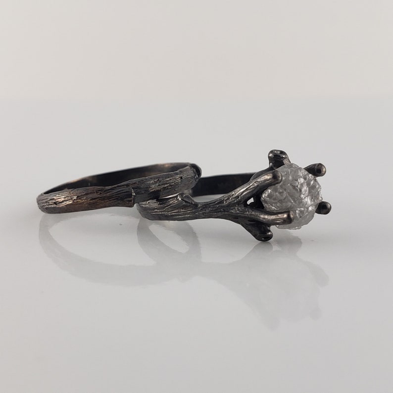 Puede incluir: Dos anillos de plata. Uno presenta un diamante en bruto de color gris claro sujeto por un engaste en forma de ramita. El otro anillo tiene una banda texturizada. Ambos anillos est&aacute;n sobre un fondo blanco.