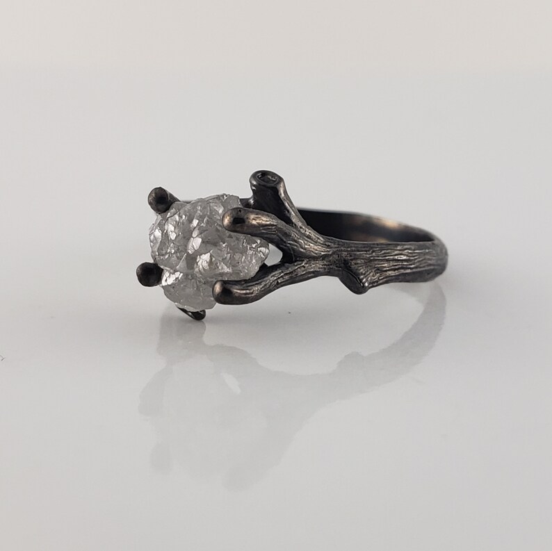 Puede incluir: Anillo plateado con una gema transparente en bruto, sujeta por un engaste en forma de ramita. La banda y el engaste del anillo tienen un acabado texturizado y oxidado, creando una est&eacute;tica r&uacute;stica y natural. La gema es el punto focal.