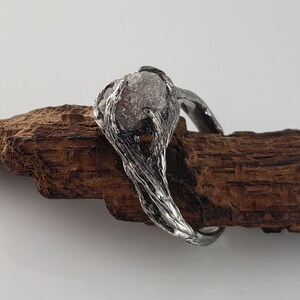 Op de afbeelding: Een zilverkleurige ring met een ruwe, lichtgekleurde edelsteen. De ring heeft een getextureerd, organisch ontwerp, dat lijkt op verweven takken. De edelsteen staat centraal, wat de ring een unieke en natuurlijke uitstraling geeft.