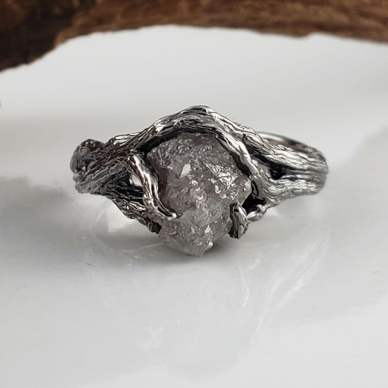 Op de afbeelding: Een zilverkleurige ring met een ruwe, lichtgrijze edelsteen. De ringband is ingewikkeld ontworpen om verweven takken na te bootsen, wat een natuurlijke, organische uitstraling cre&euml;ert. De edelsteen is het middelpunt, stevig vastgehouden in de takachtige zetting.