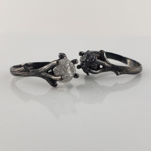 Puede incluir: Dos anillos de plata con un dise&ntilde;o similar a una ramita. Un anillo presenta una piedra clara y en bruto, mientras que el otro tiene una piedra oscura y texturizada. Los anillos se colocan sobre un fondo blanco, mostrando su est&eacute;tica &uacute;nica inspirada en la naturaleza.